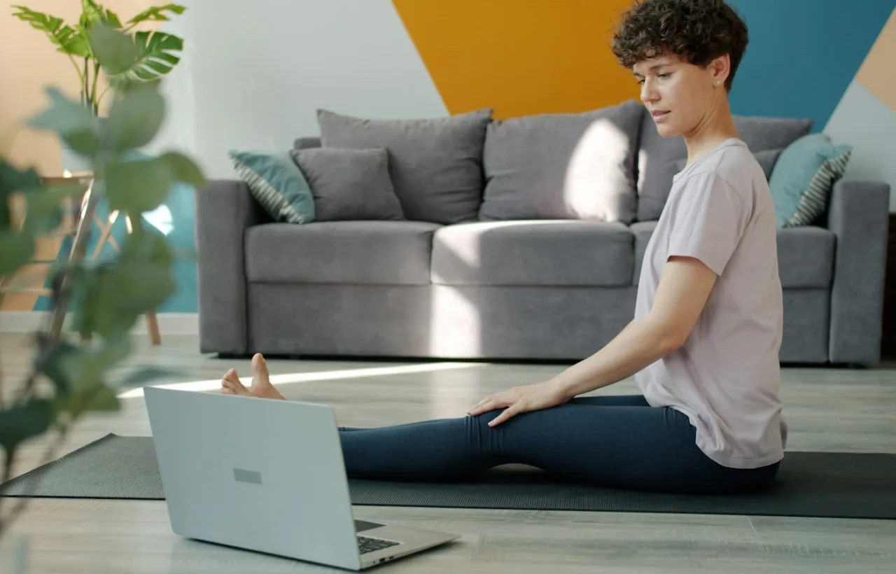 Woman following a home workout video on her laptop while stretching on a yoga mat